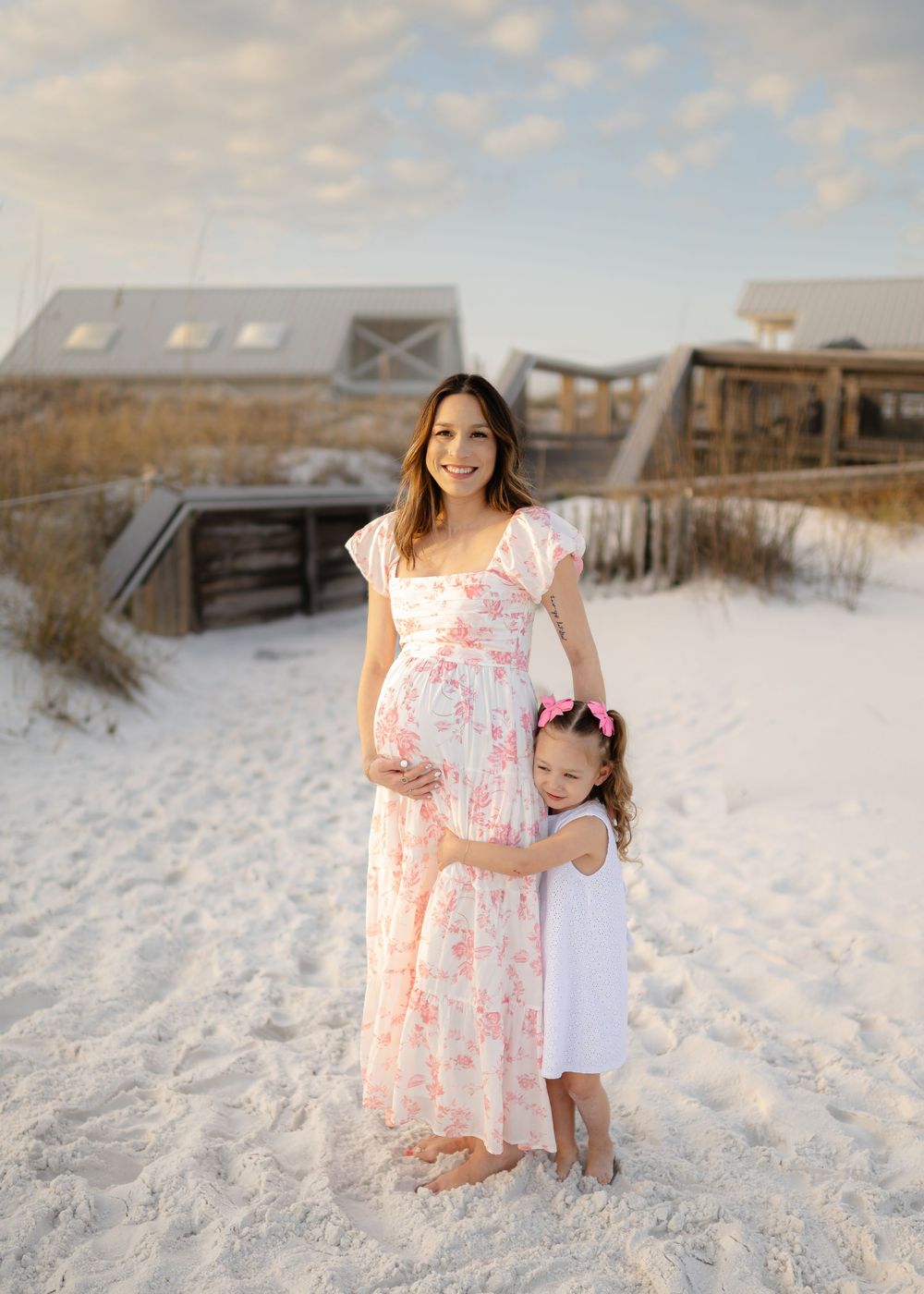 Pregnant woman in floral dress with young girl on white sand beach at sunset. Beautiful sunset photos with a professional photographer.
