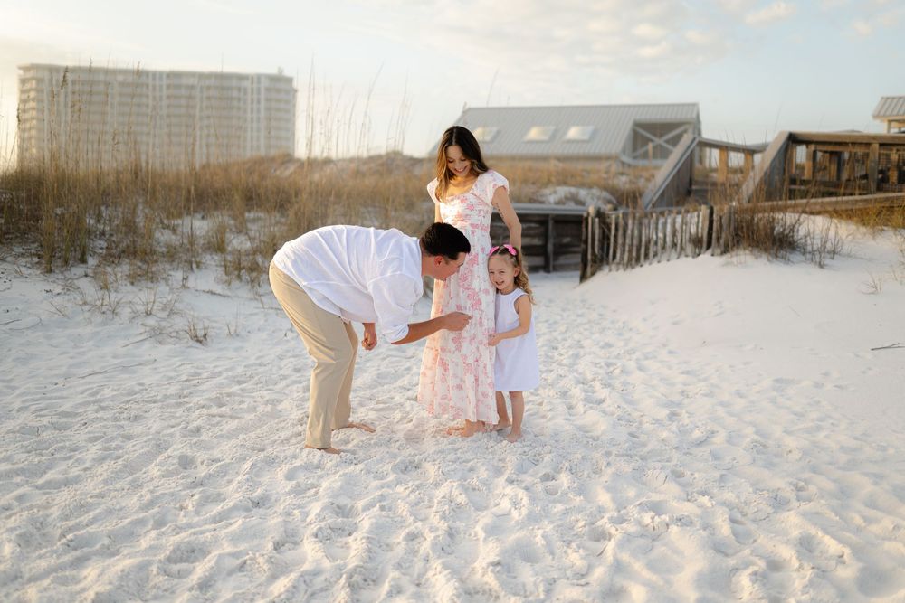 Family of three enjoying a sunny day on a white sand beach, with a father bending down to his young daughter.