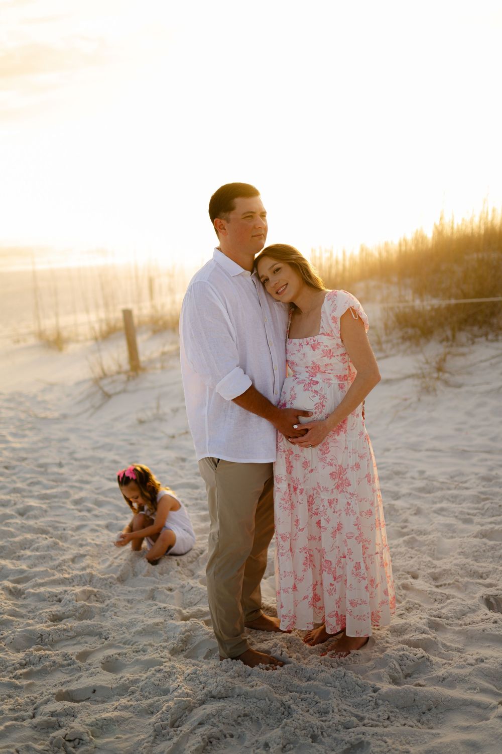 Pregnant couple embracing on sandy beach at golden sunset, with young child playing in sand behind them.