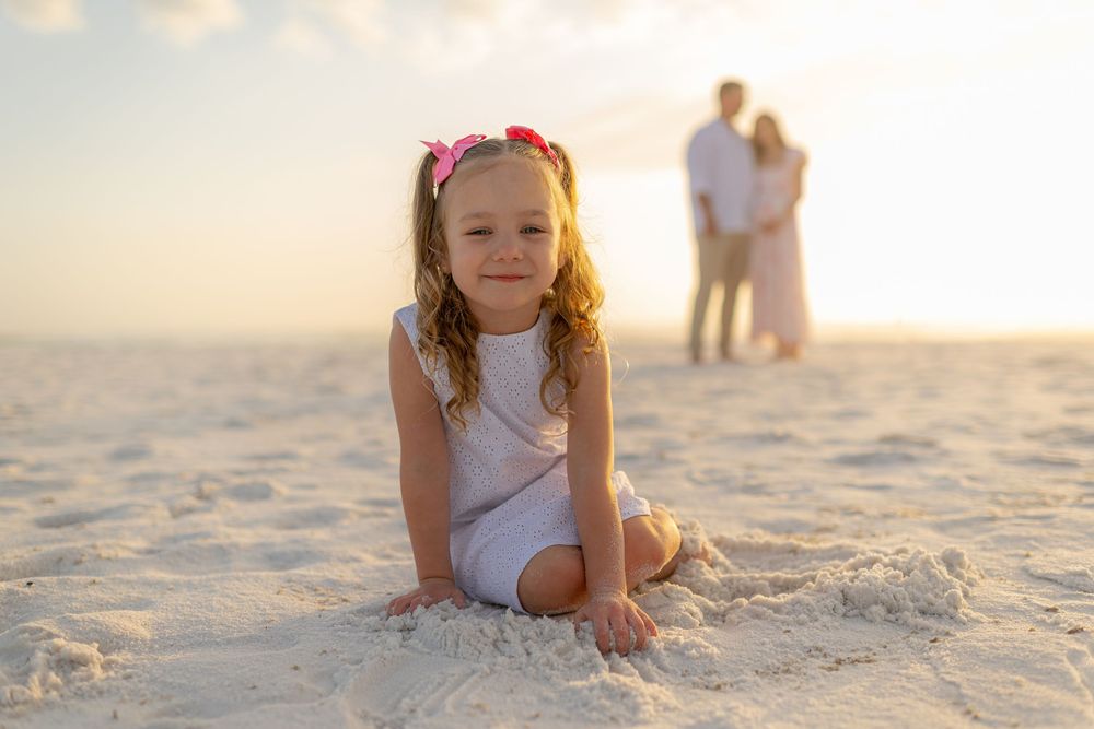 A young girl in a white outfit sits smiling on a sandy beach at sunset, with a couple embracing in the background.
