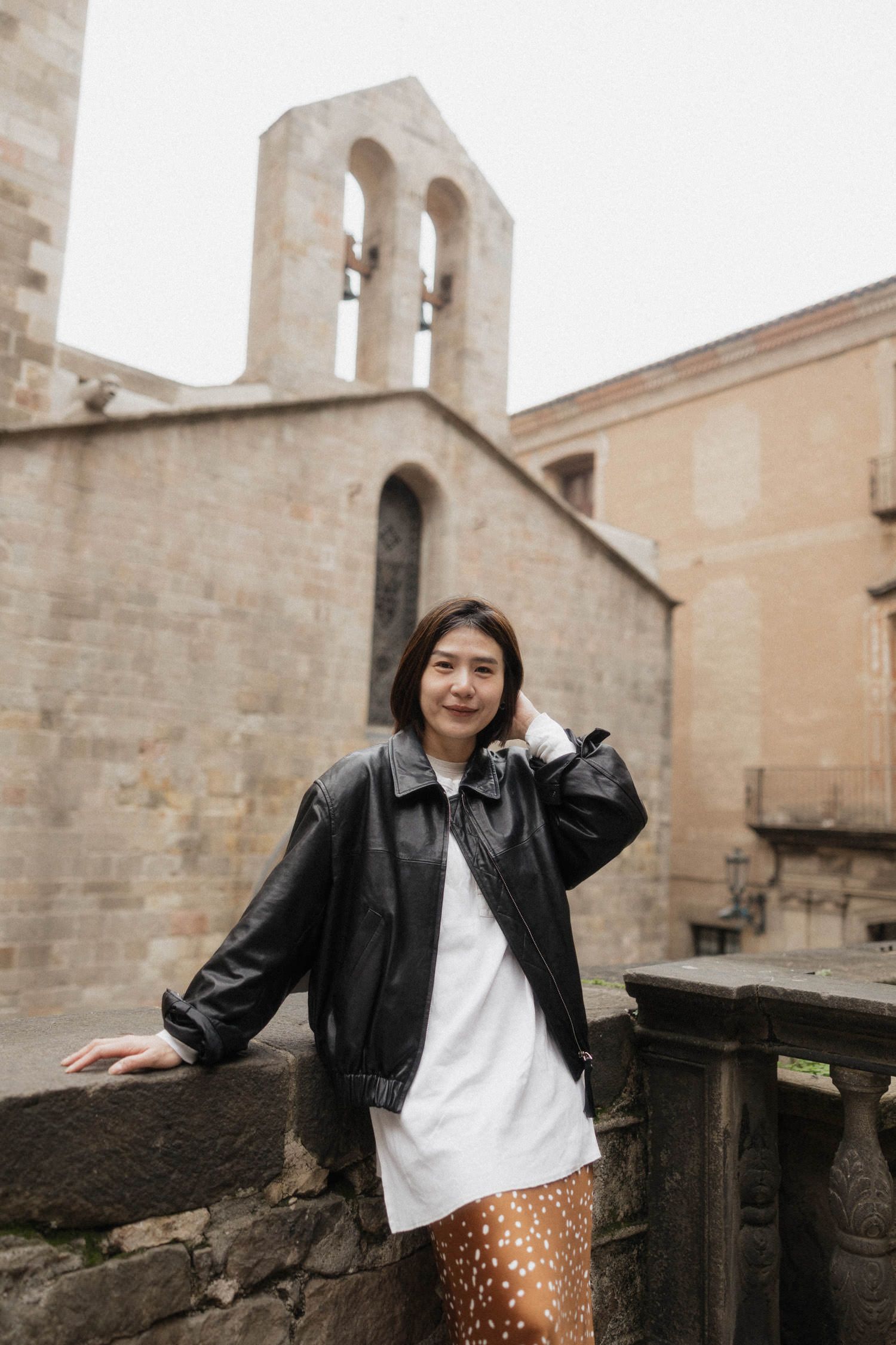 Young woman in black leather jacket and white dress poses by stone wall near historic church bell tower.