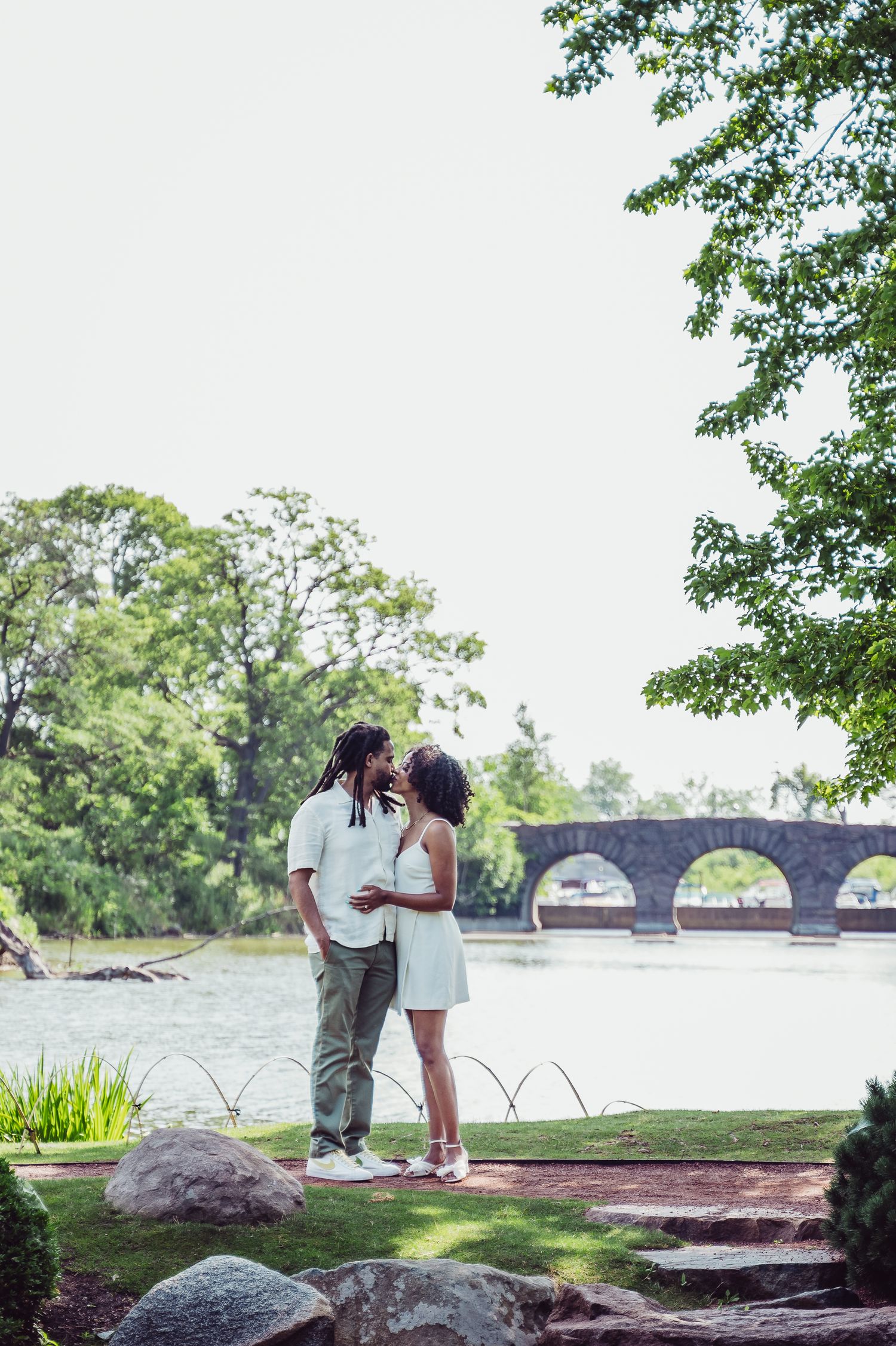 A couple shares an intimate moment by a lake with a stone bridge in the background on a sunny summer day.