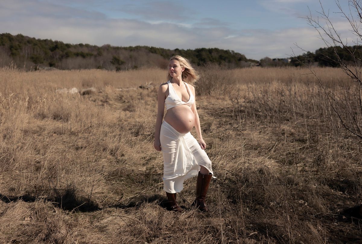 gravid kvinna står på en åker. vinden blåser i hennes hår och hon har ett vitt set på sig och bruna boots.