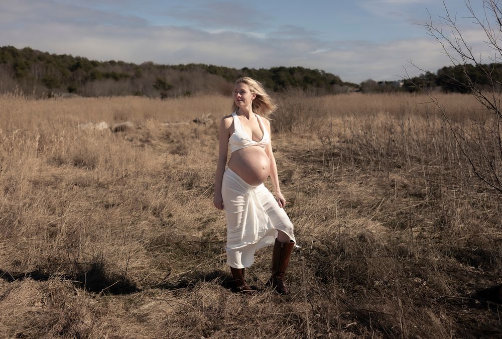 gravid kvinna står på en åker. vinden blåser i hennes hår och hon har ett vitt set på sig och bruna boots.