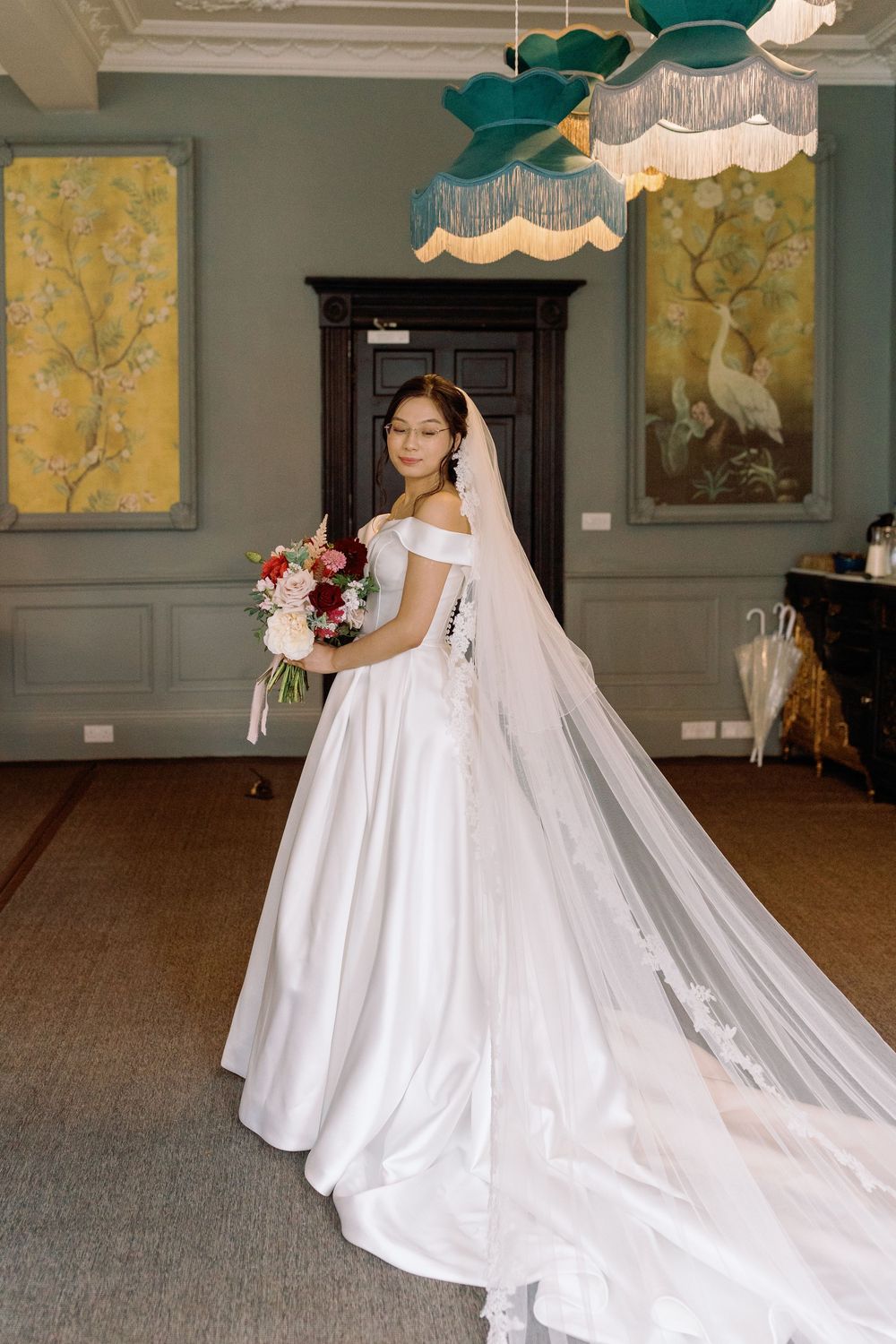 A bride in an elegant off-shoulder white gown with long train poses against sage green walls with floral artwork.