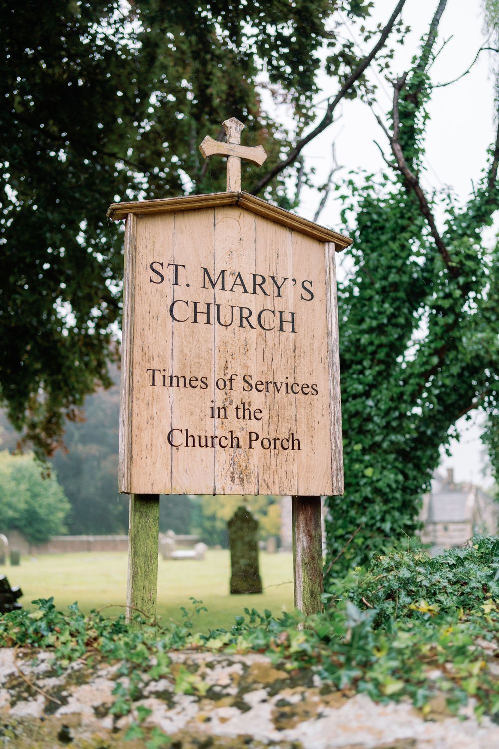 Wooden church sign for St. Mary's displaying 'Times of Services' and 'Church Porch' with a cross on top.