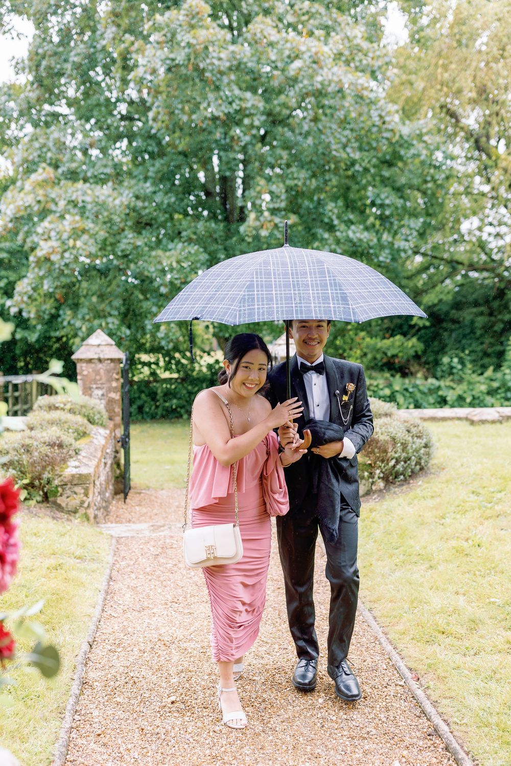 A couple shares an intimate moment under a checkered umbrella on a garden path surrounded by lush greenery taken by Maddie Waters