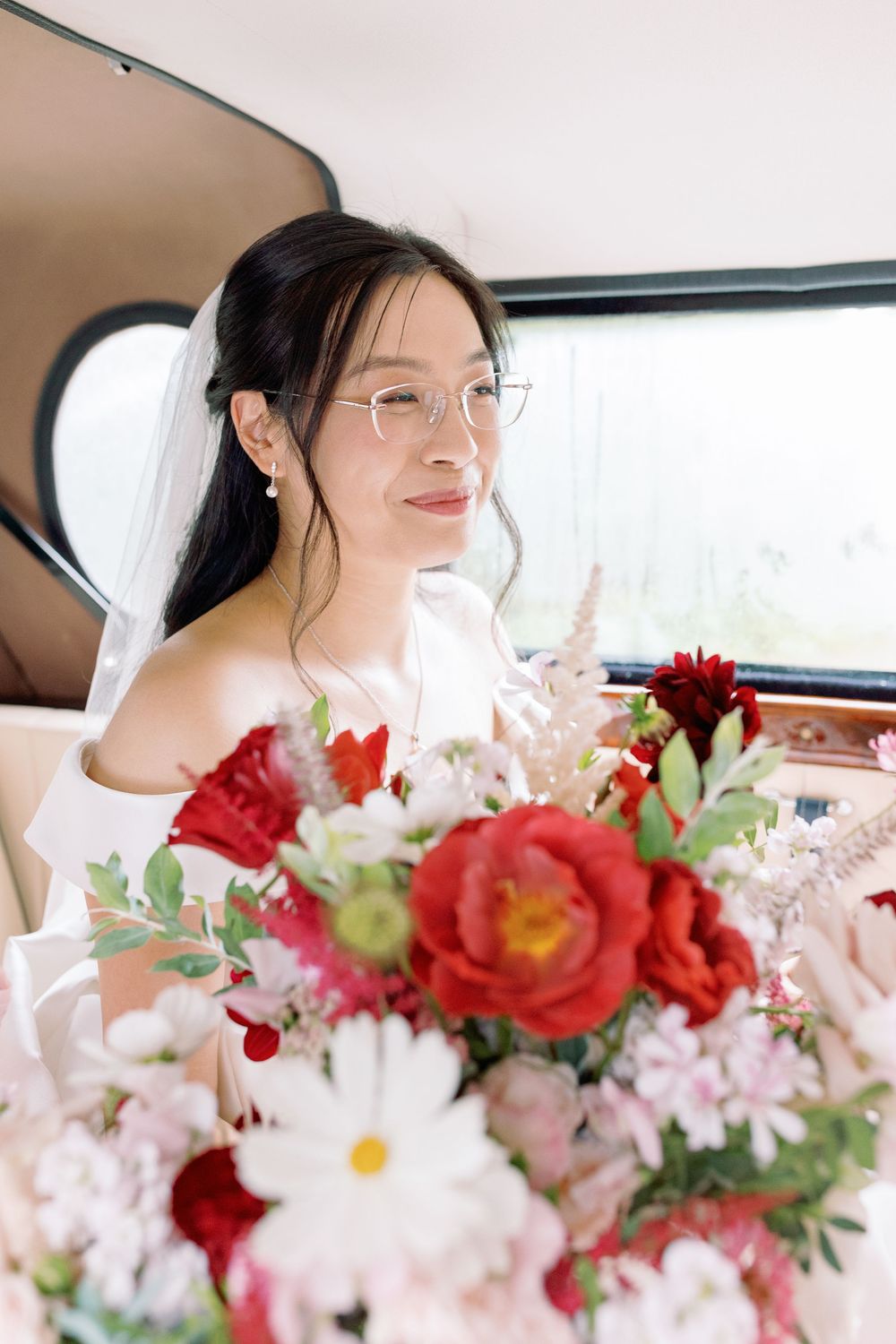 A bride in a vintage car holds a vibrant bouquet of red and white flowers taken by Maddie Waters Photography