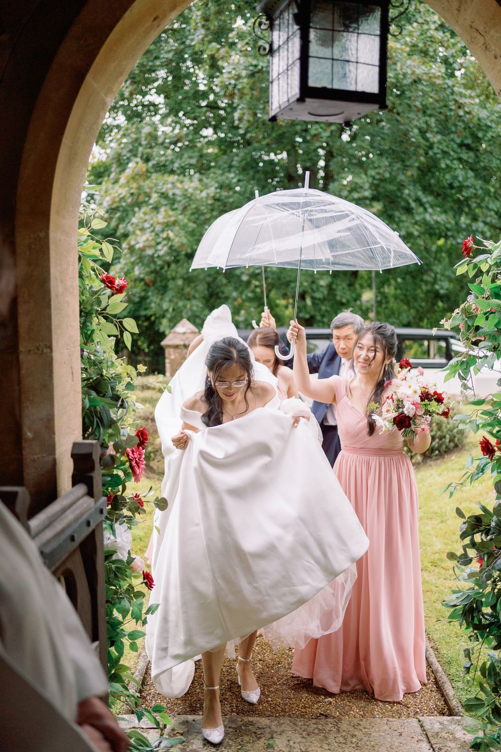 Wedding party walks with clear umbrellas under an outdoor archway decorated with pink roses taken by Maddie Waters Photography