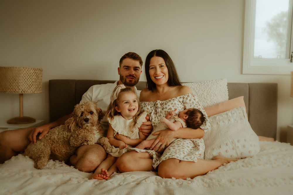 Picnic Point Newborn Photography session as a family relaxes together on a cozy bed with their goldendoodle dog in a warmly lit bedroom.