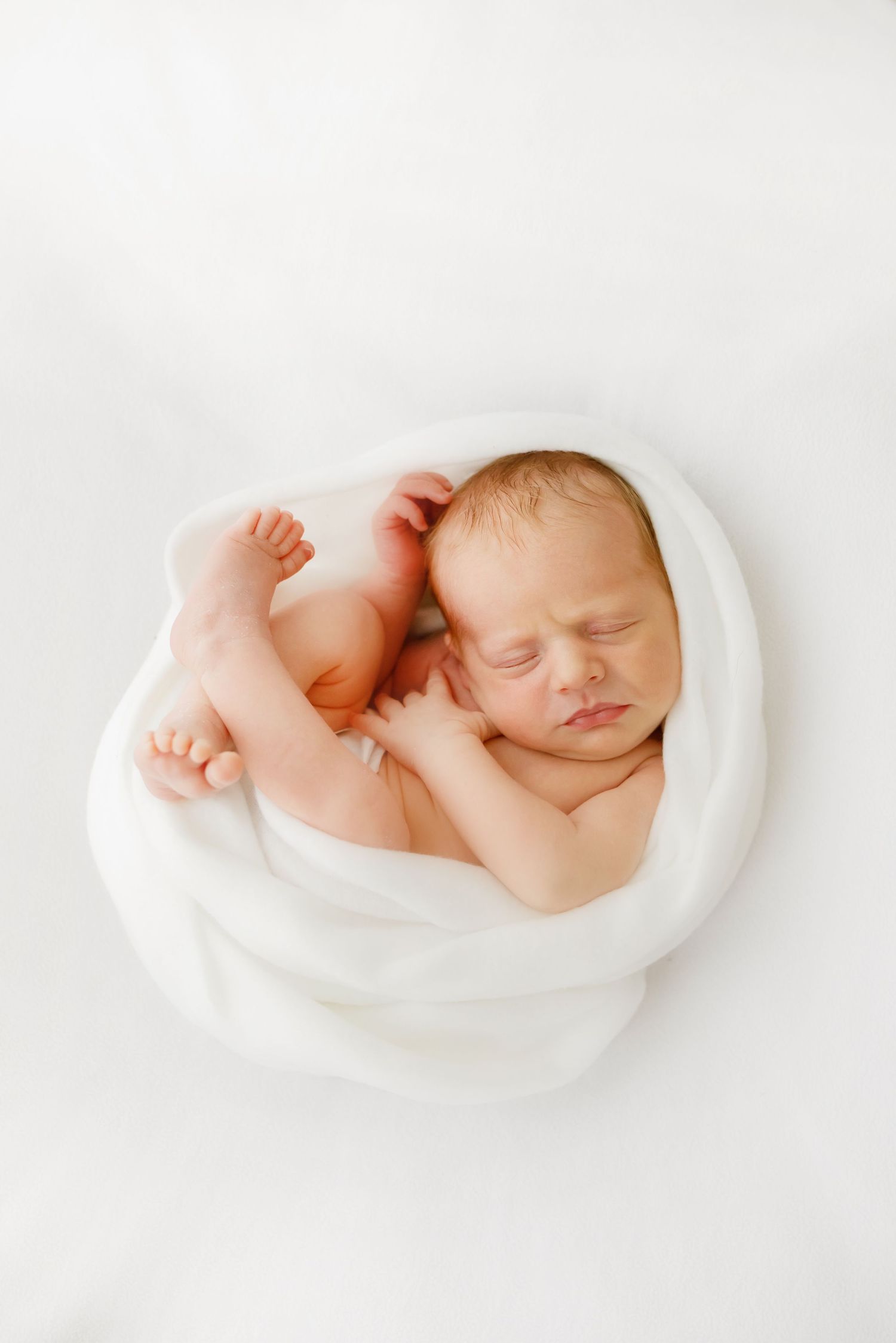 En sovande nyfödd bebis hopkrupen lugnt på mjukt vitt tyg i en minimalistisk komposition.