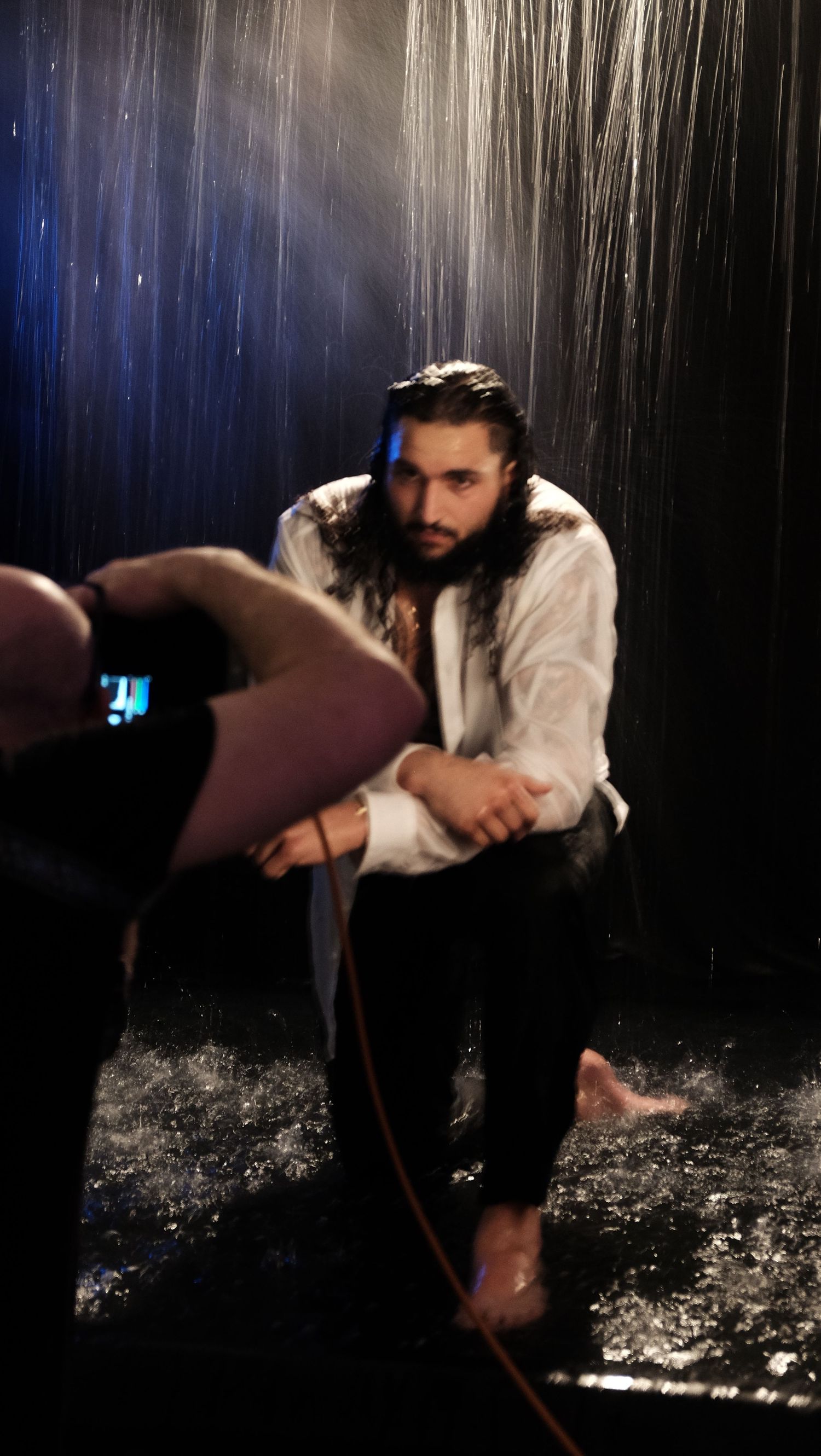 Dramatic photo shoot in the rain with moody lighting showing a seated figure in white clothing against dark background.