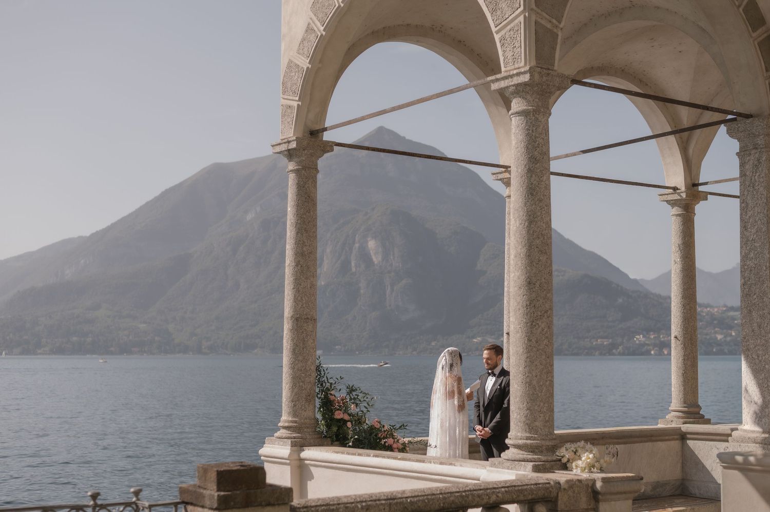 A couple stands under an elegant stone archway overlooking a scenic Mediterranean coastline with mountains in the distance.