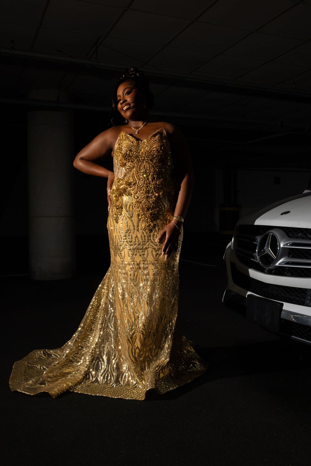 A person poses in a stunning gold sequined evening gown with a mermaid silhouette against a dark backdrop with a car visible.