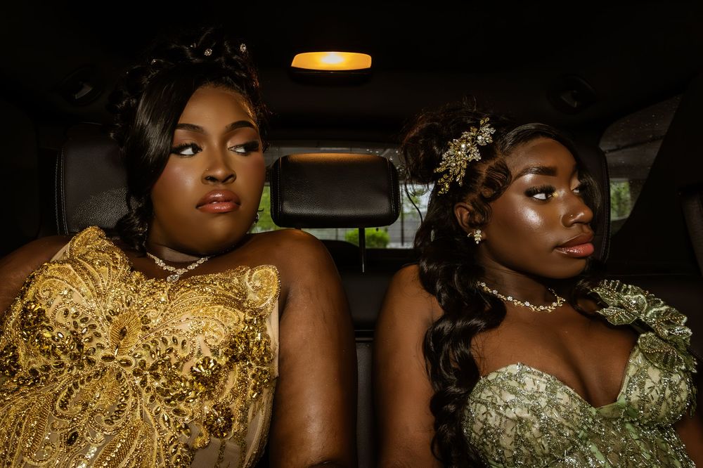 Two people wearing elegant gold and silver formal gowns sit together in what appears to be the back of a vehicle.