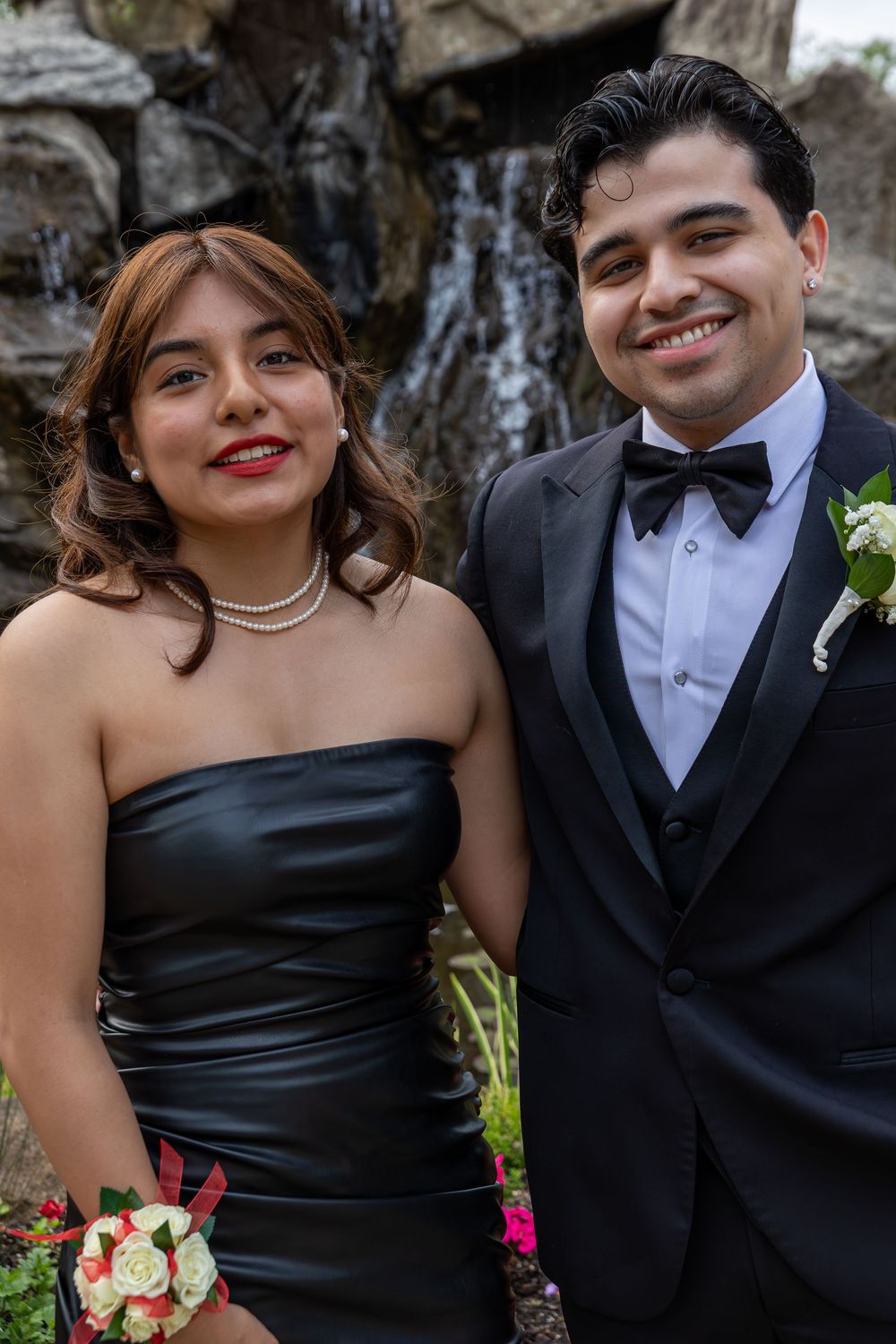 A couple in formal attire poses in front of a waterfall backdrop, with the woman in a black dress holding a bouquet.