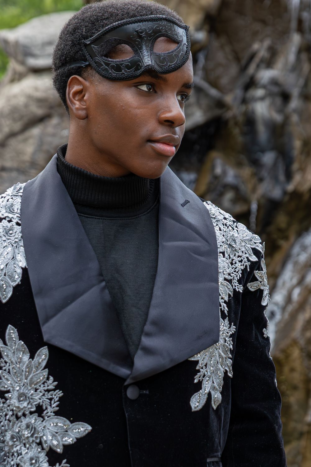 A person in a black and silver floral jacket and steampunk goggles poses against a rocky backdrop.
