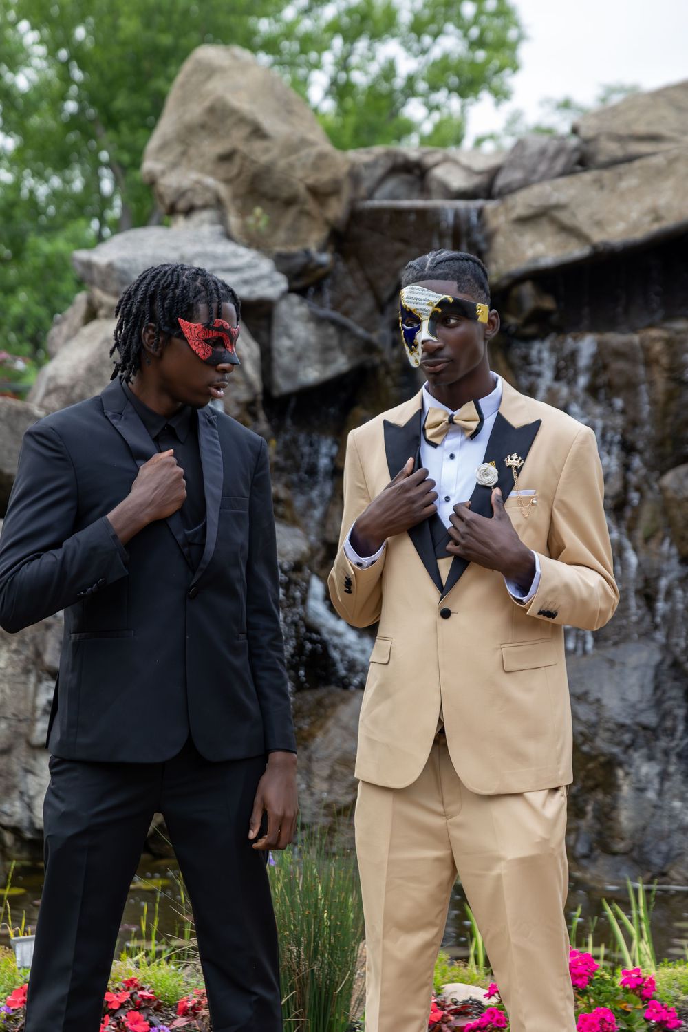 Two friends in formal suits strike playful poses in front of a rock wall with a waterfall.