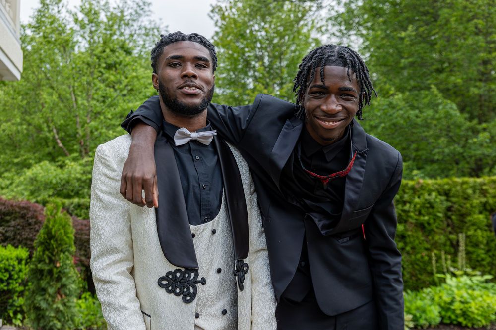 Two people in formal attire smile and embrace while standing in a lush green outdoor setting.