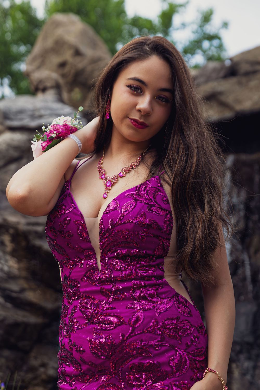 A woman in a sparkly magenta gown poses elegantly against a rocky backdrop with flowers.