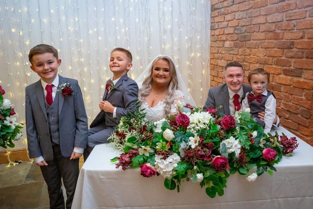 Bride and groom pose with three boys in matching grey suits at a decorated wedding table with burgundy floral arrangements.