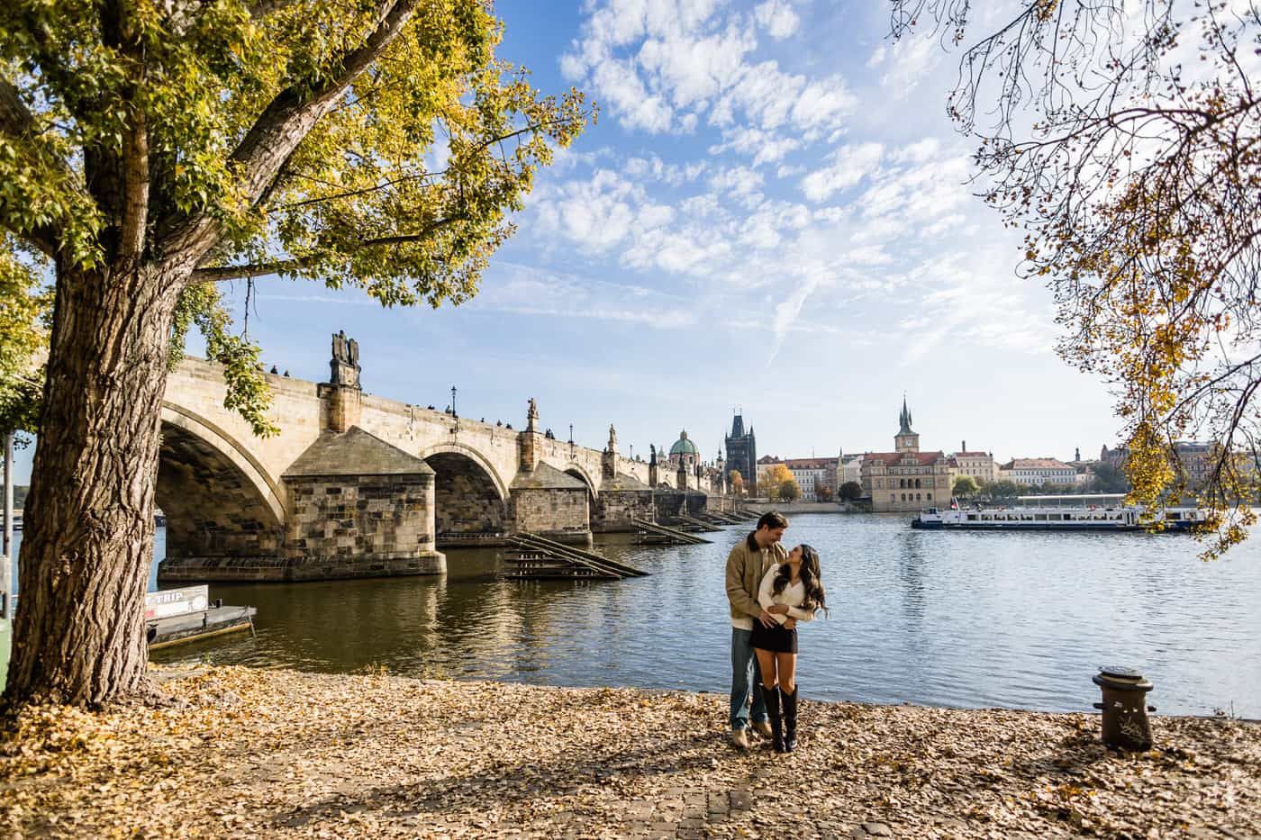 Romantic couple embraces on a scenic overlook with historic European church architecture and ornate railings in background.
