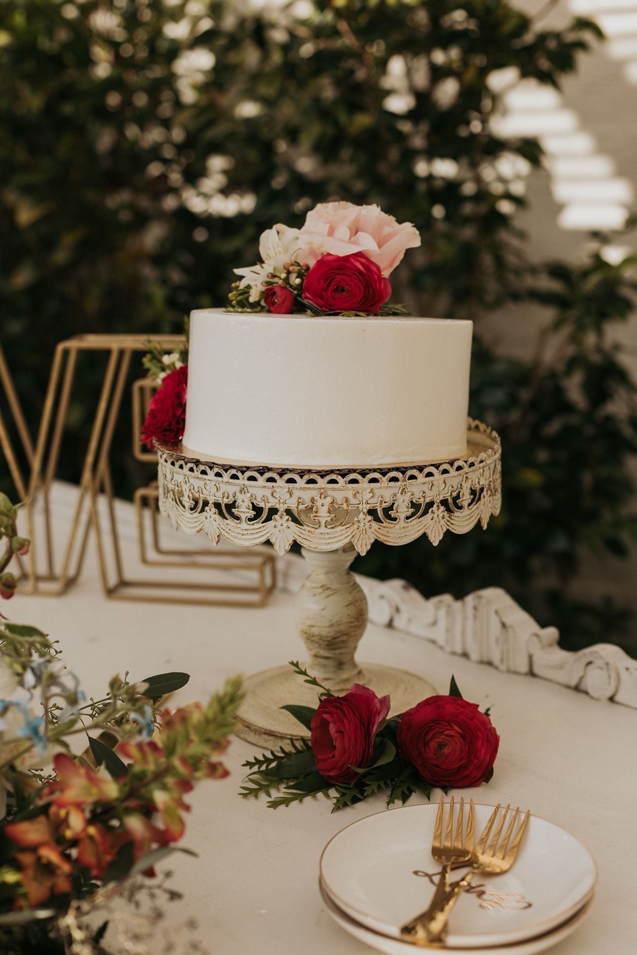 Elegant two-tier white wedding cake topped with red and pink roses on a ornate cake stand with gold forks and floral decor.