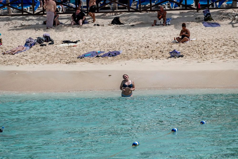 Tourists relaxing on a white sandy Caribbean beach with crystal clear turquoise water and blue buoy markers.