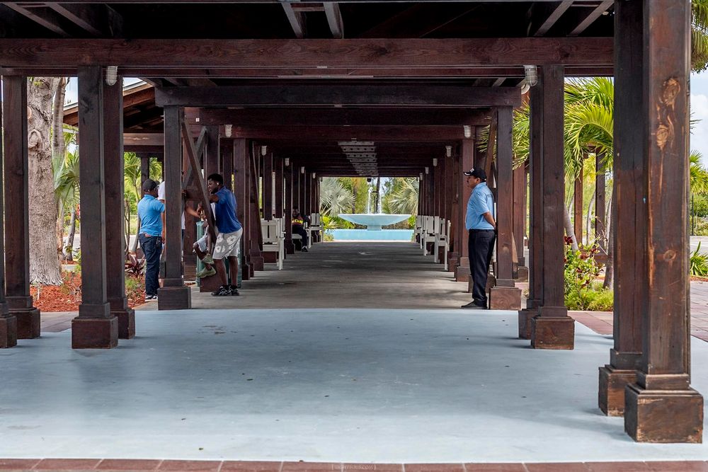 Covered wooden pergola walkway with visitors strolling toward a tropical pool area surrounded by palm trees.