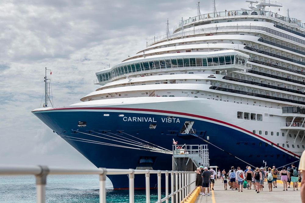 Carnival Vista cruise ship docked at a tropical port with passengers walking along the pier on a cloudy day.