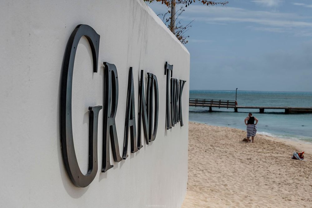 Grand Turk sign on a white wall with a sandy beach, pier, and turquoise ocean in the background.
