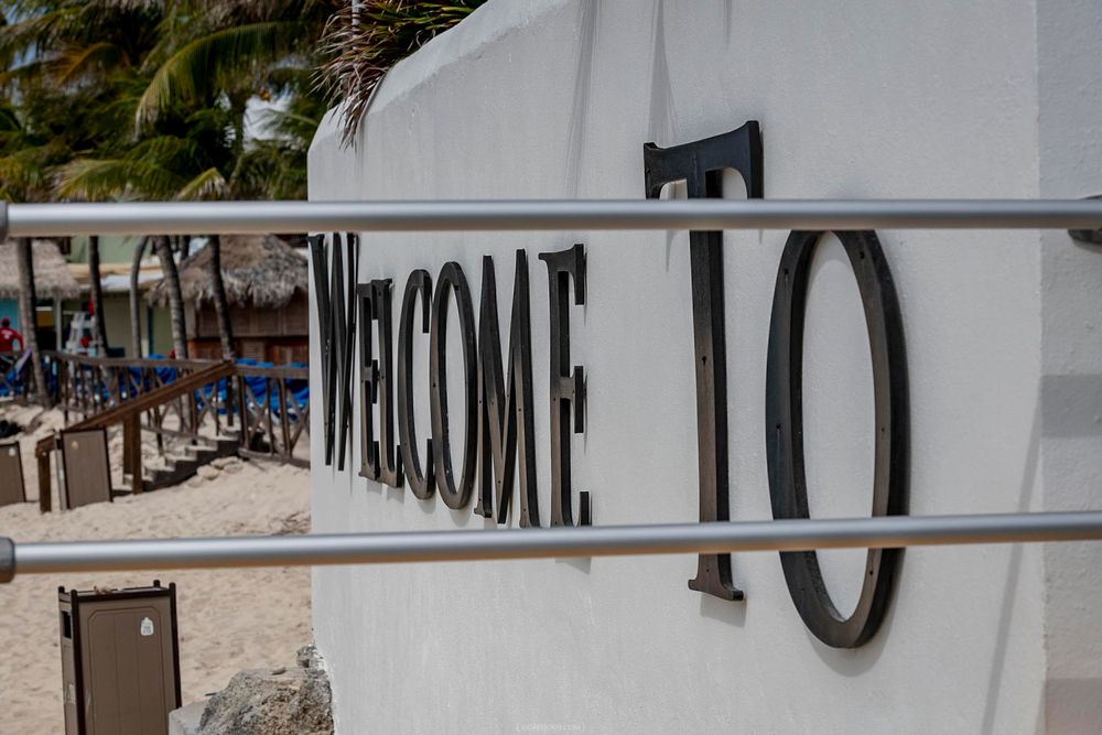Welcome To sign on a white wall at a tropical beach resort with palm trees and lounge chairs.