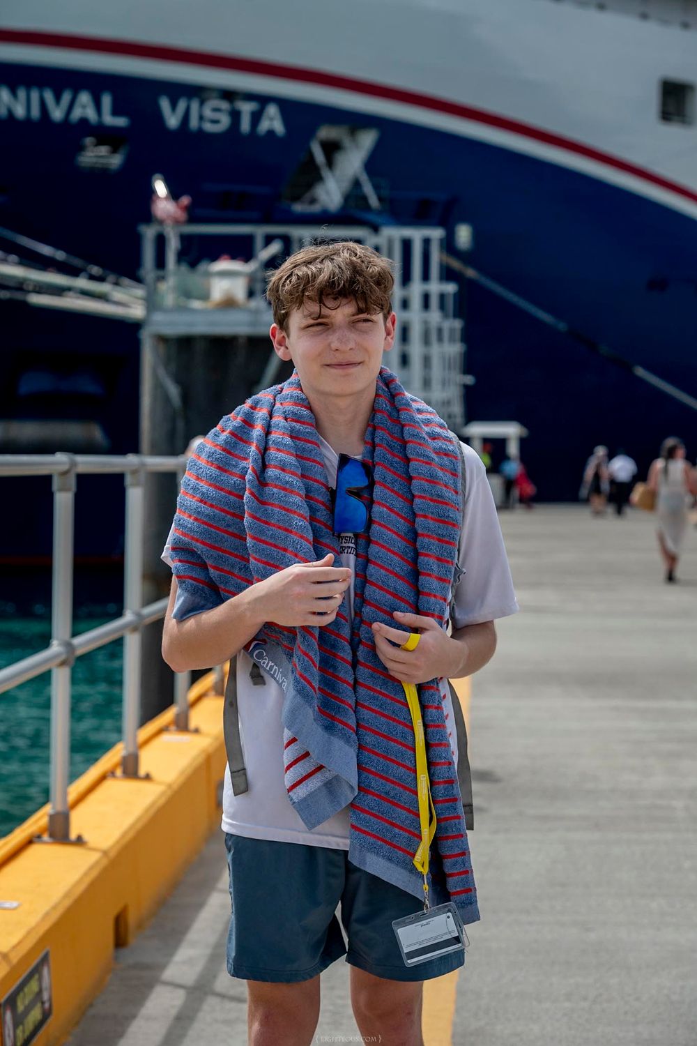 Teen boy with striped towel stands on dock in front of Carnival Vista cruise ship on sunny day.