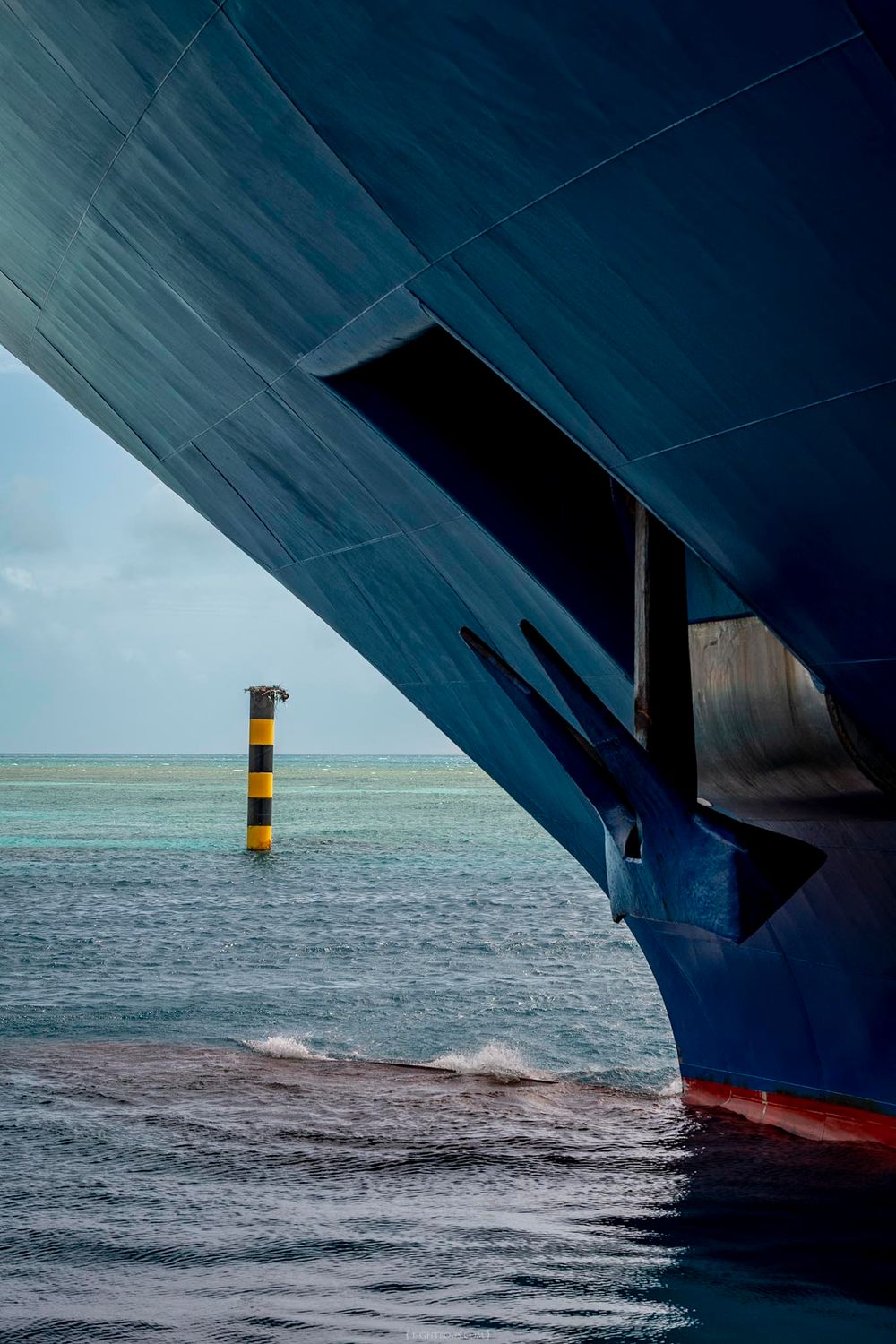 View of a yellow and black channel marker through the bow and anchor of a large blue cargo ship at sea.