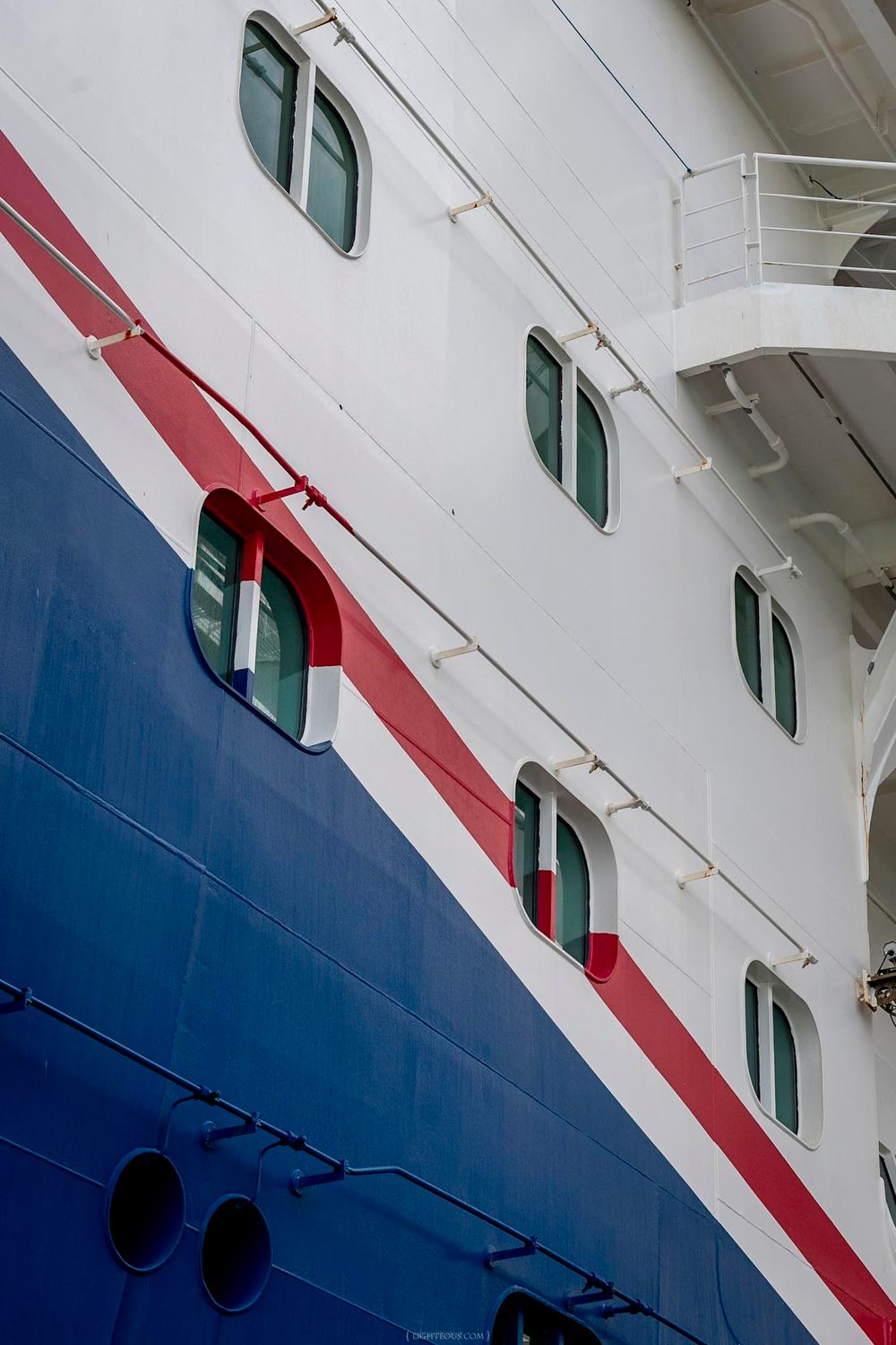 Close-up of a cruise ship hull featuring white upper decks with green windows and a bold red and blue stripe.
