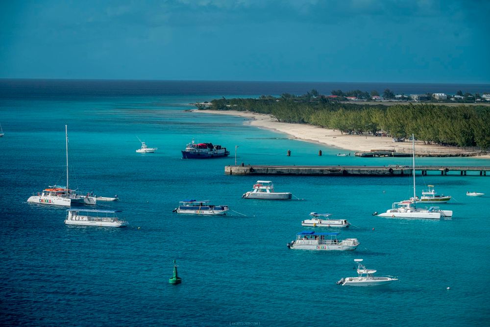 Aerial view of turquoise Caribbean waters with sailboats and motorboats anchored near a tropical island pier.