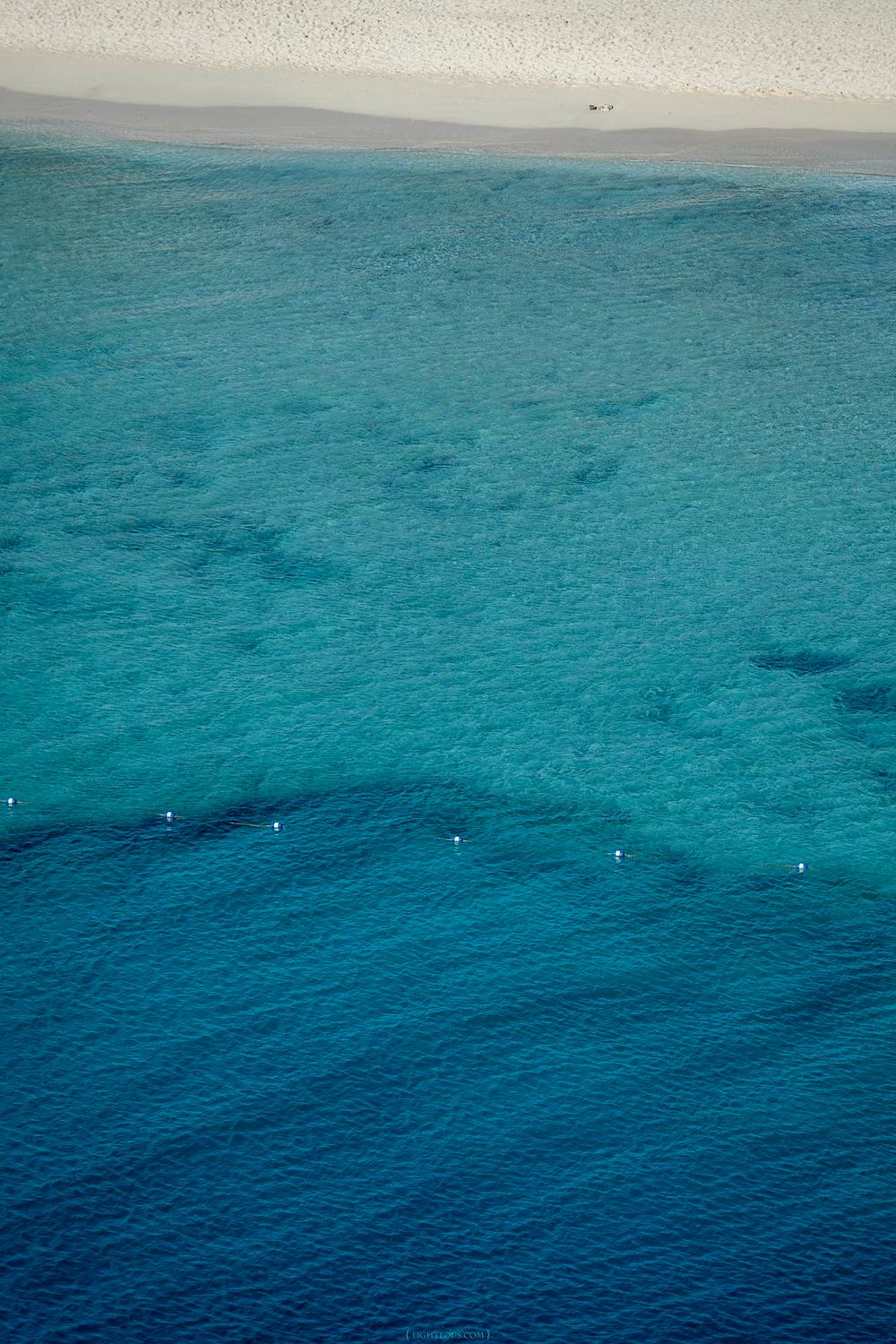Aerial view of crystal clear turquoise ocean water with a white sandy beach shoreline in the background.
