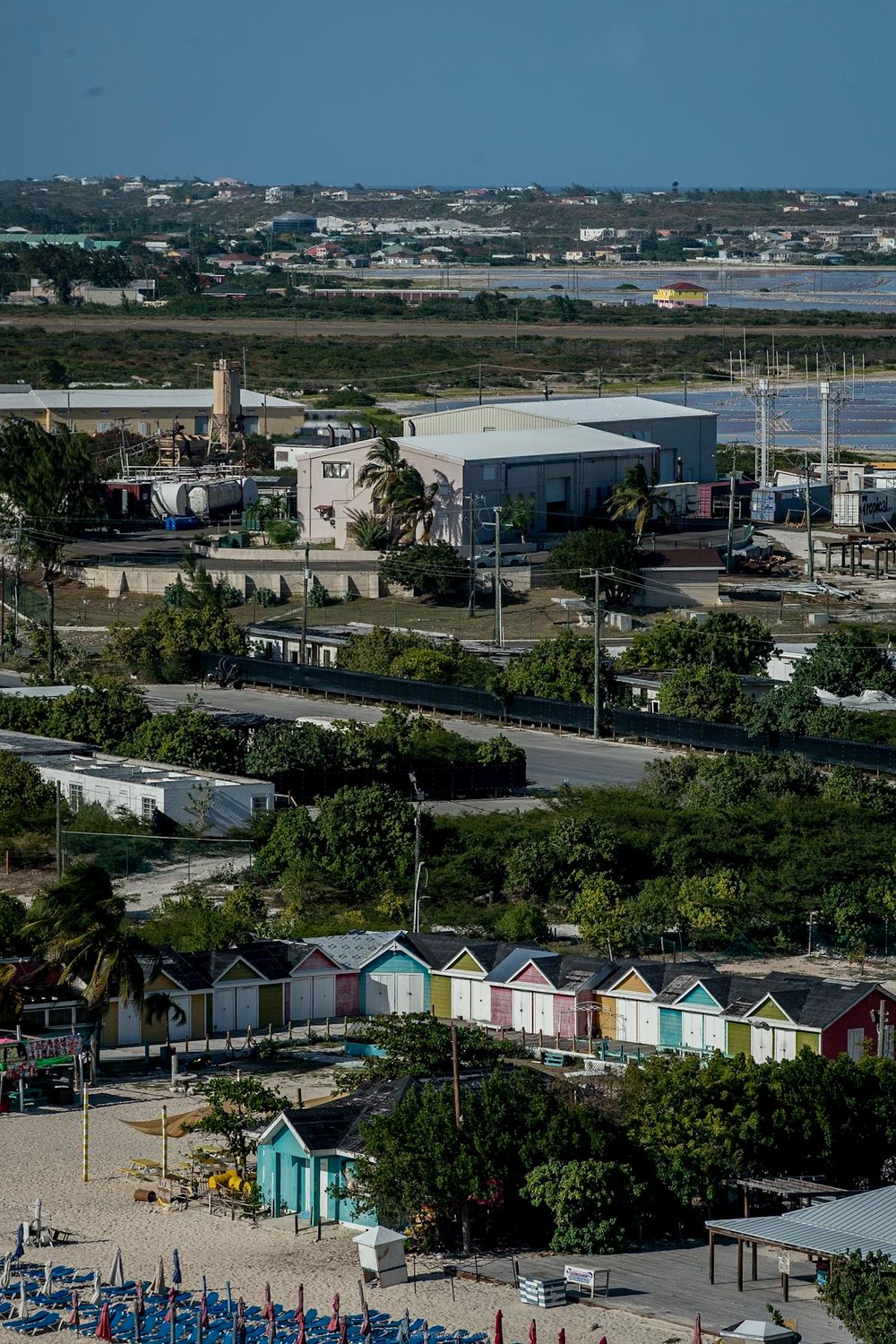 Colorful beach huts line a sandy shore near an industrial area and road in a tropical coastal town.