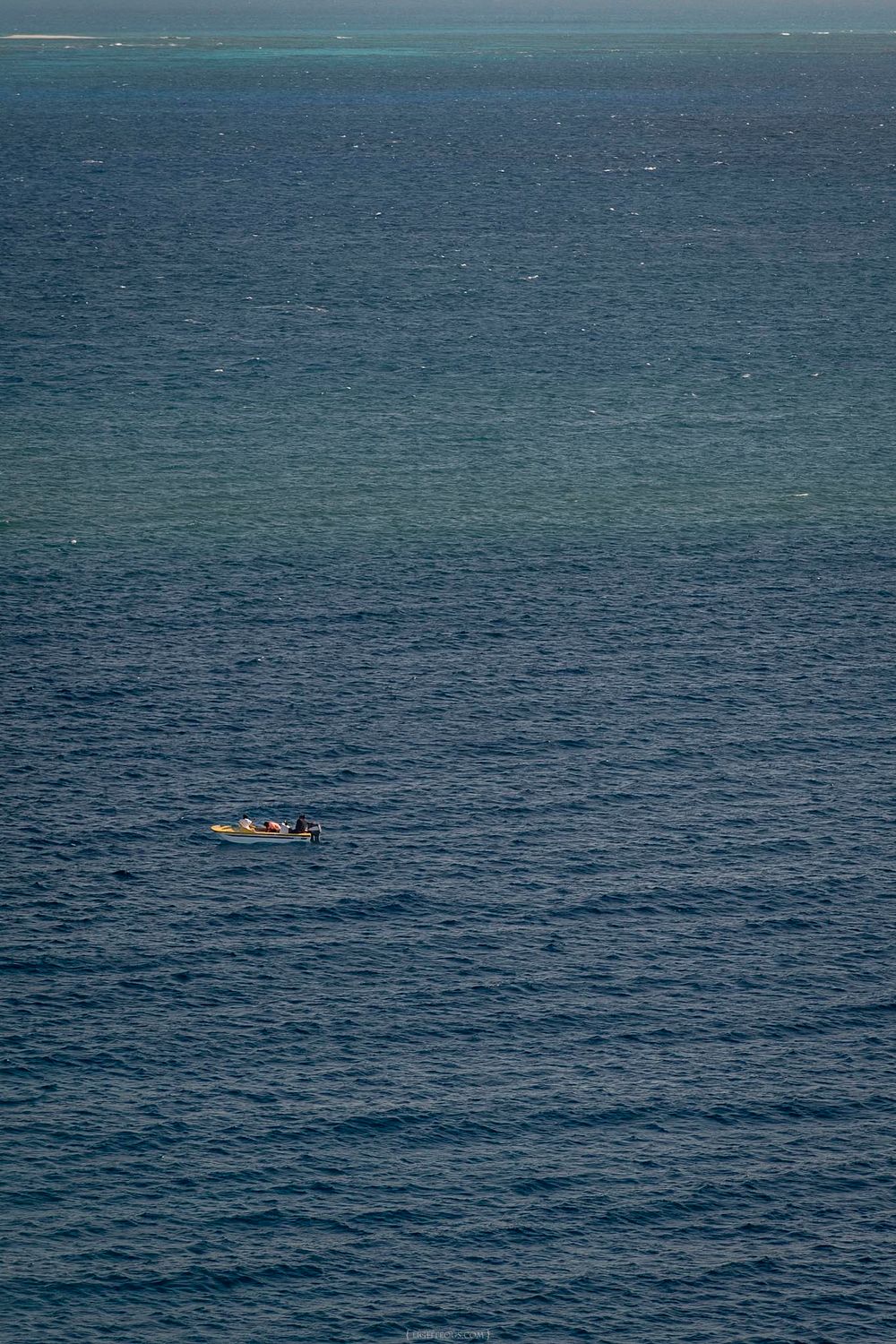 Lone small boat floating on calm deep blue ocean waters with a distant reef visible on the horizon.