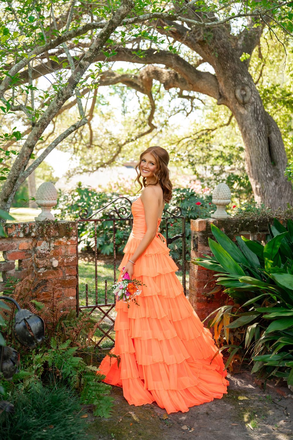 Young woman in a tiered coral orange gown holds a bouquet in a lush garden with iron gates and trees.