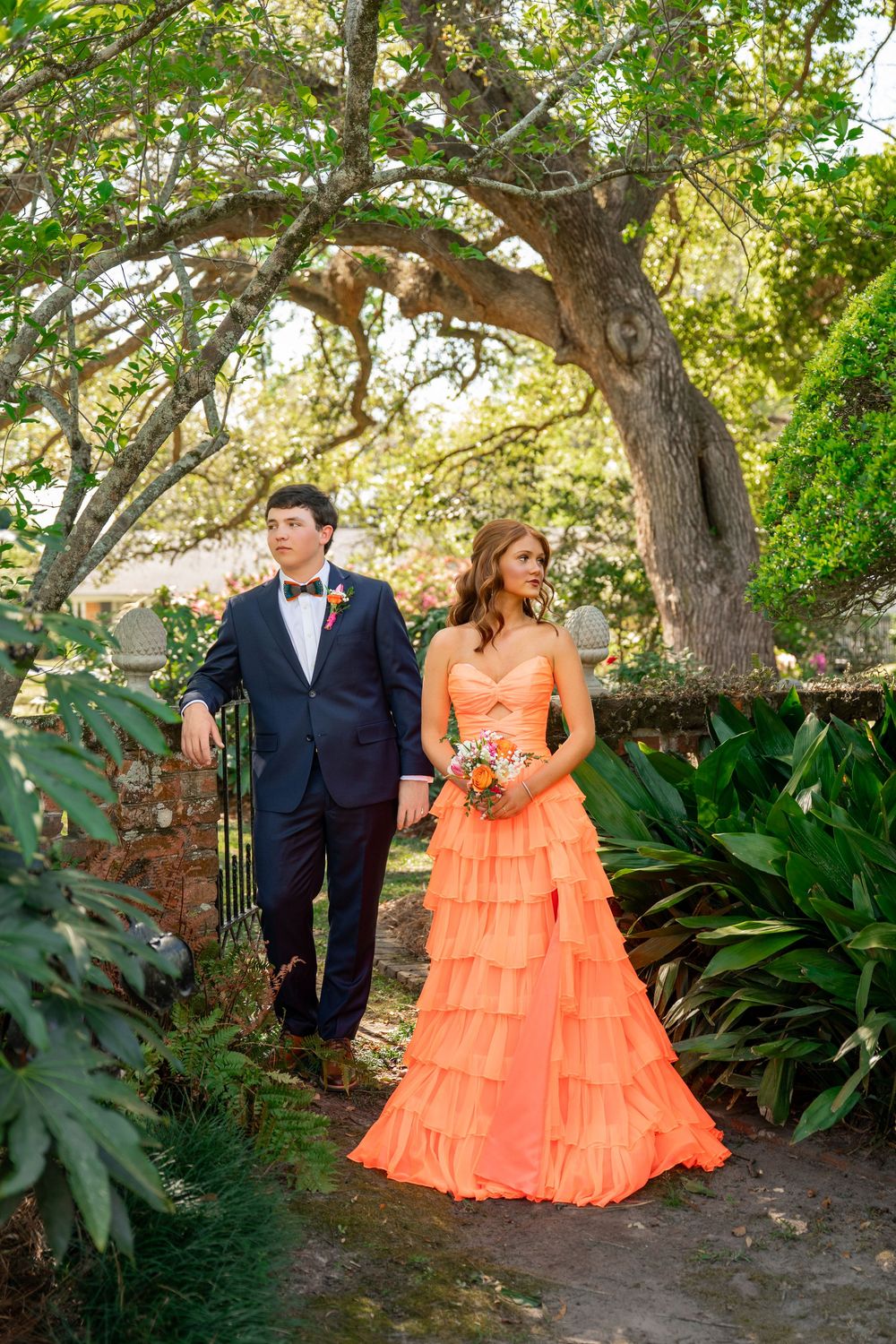 Couple in formal wear poses under oak tree, woman in orange tiered gown holding bouquet, man in navy suit with bow tie.