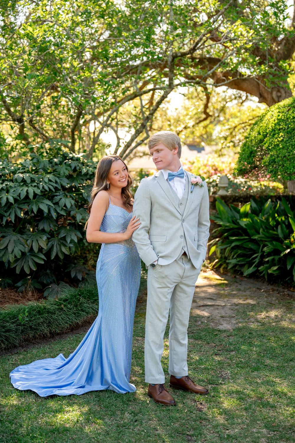 A couple in prom attire poses in a lush garden, she in a blue gown and he in a tan suit with bow tie.