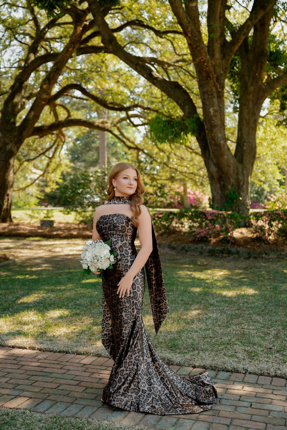 Young woman in leopard print strapless gown holding white bouquet poses on brick path in lush garden setting.
