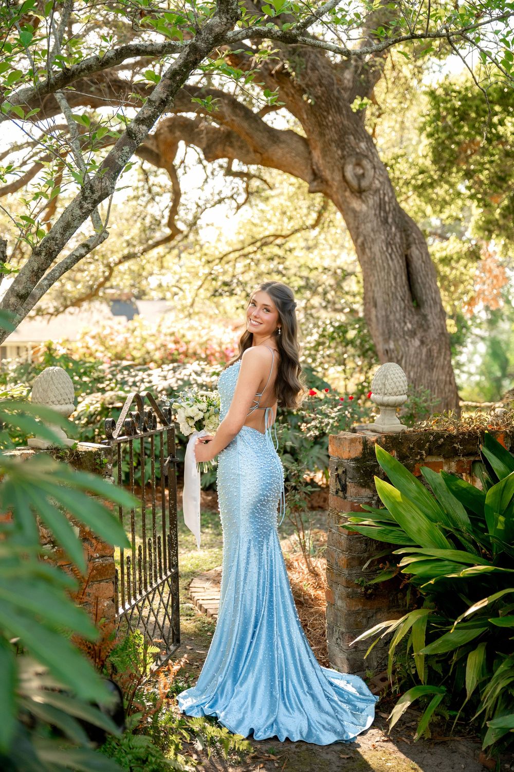 Young woman in a light blue sequin mermaid gown posing in a lush garden with a large oak tree.
