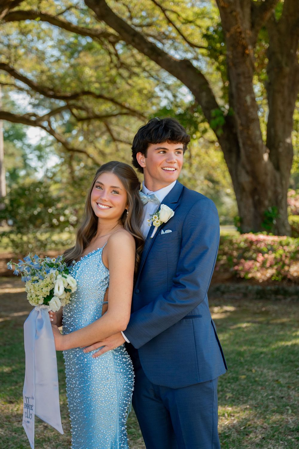 A smiling couple dressed in prom attire, blue gown and navy suit, posing outdoors under a large tree.