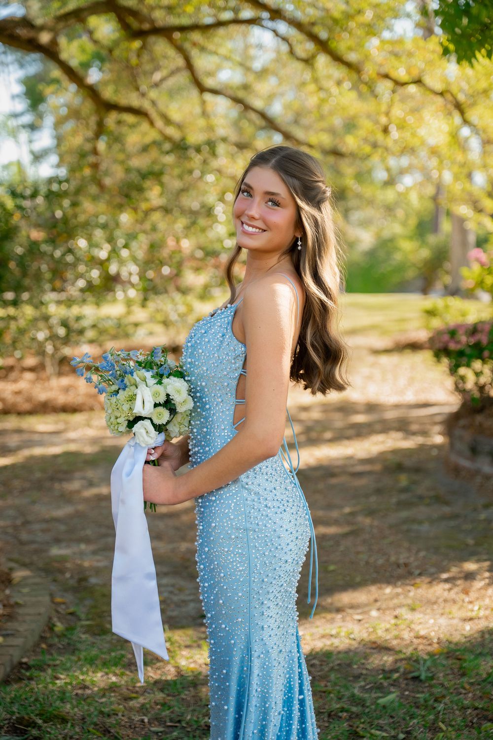 Young woman in elegant blue beaded mermaid prom dress holding white floral bouquet outdoors under trees.