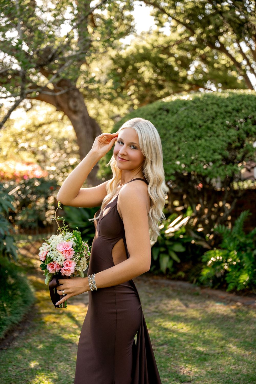 Blonde woman in brown satin gown holding pink bouquet poses in lush garden.