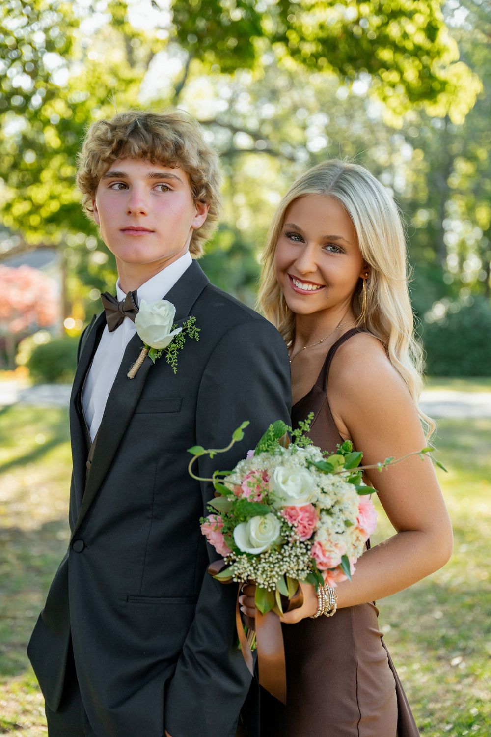 Teen prom couple in garden, boy in black tuxedo with boutonniere, girl in brown gown with bouquet.