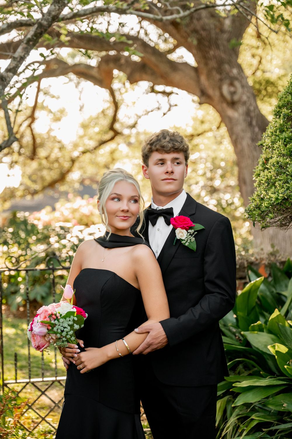 Young couple in black formal wear pose outdoors, she holds pink bouquet, he wears red rose boutonniere.