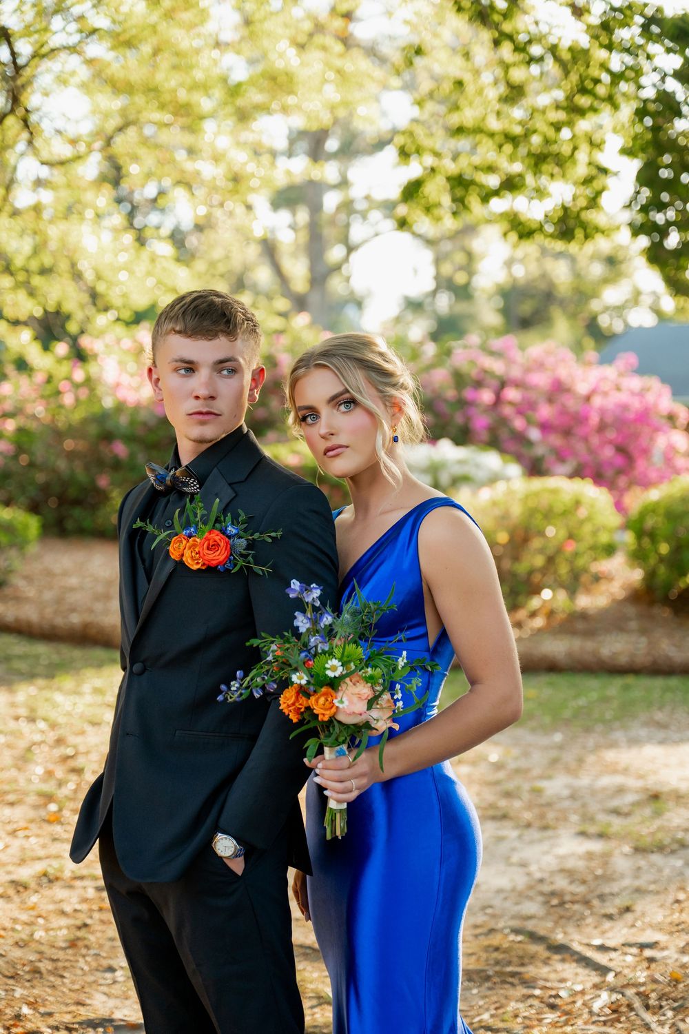 Young couple in prom attire, man in black tux with boutonniere, woman in royal blue gown holding floral bouquet outdoors.